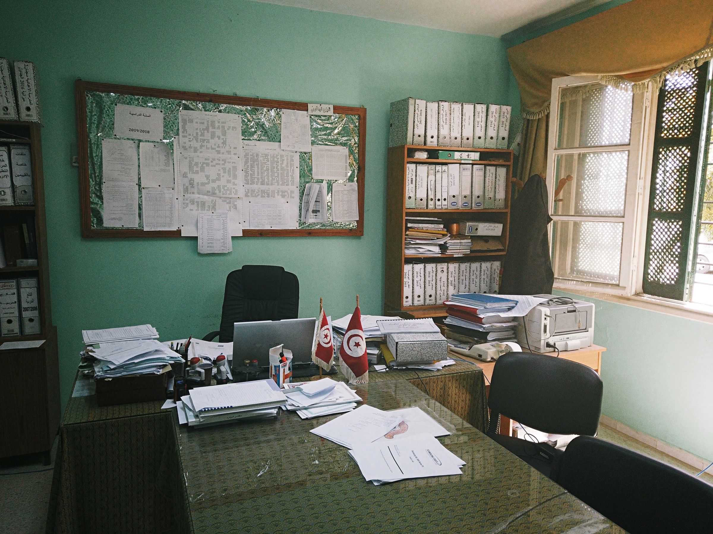 Photo d'un bureau de directeur dans une école tunisienne comportant un bureau et des chaises ainsi qu'une étagère remplie de classeurs et un tableau avec plusieurs affiches sur un mur vert pâle