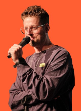 Smiling young man with short hair poses against a dark background, wearing a green button-up shirt.