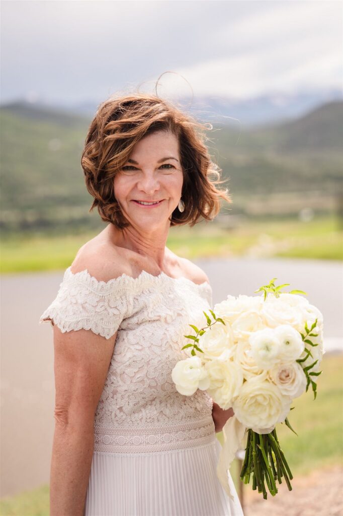 Bride in an off-the-shoulder lace dress holding a white bouquet against a softly blurred mountain landscape