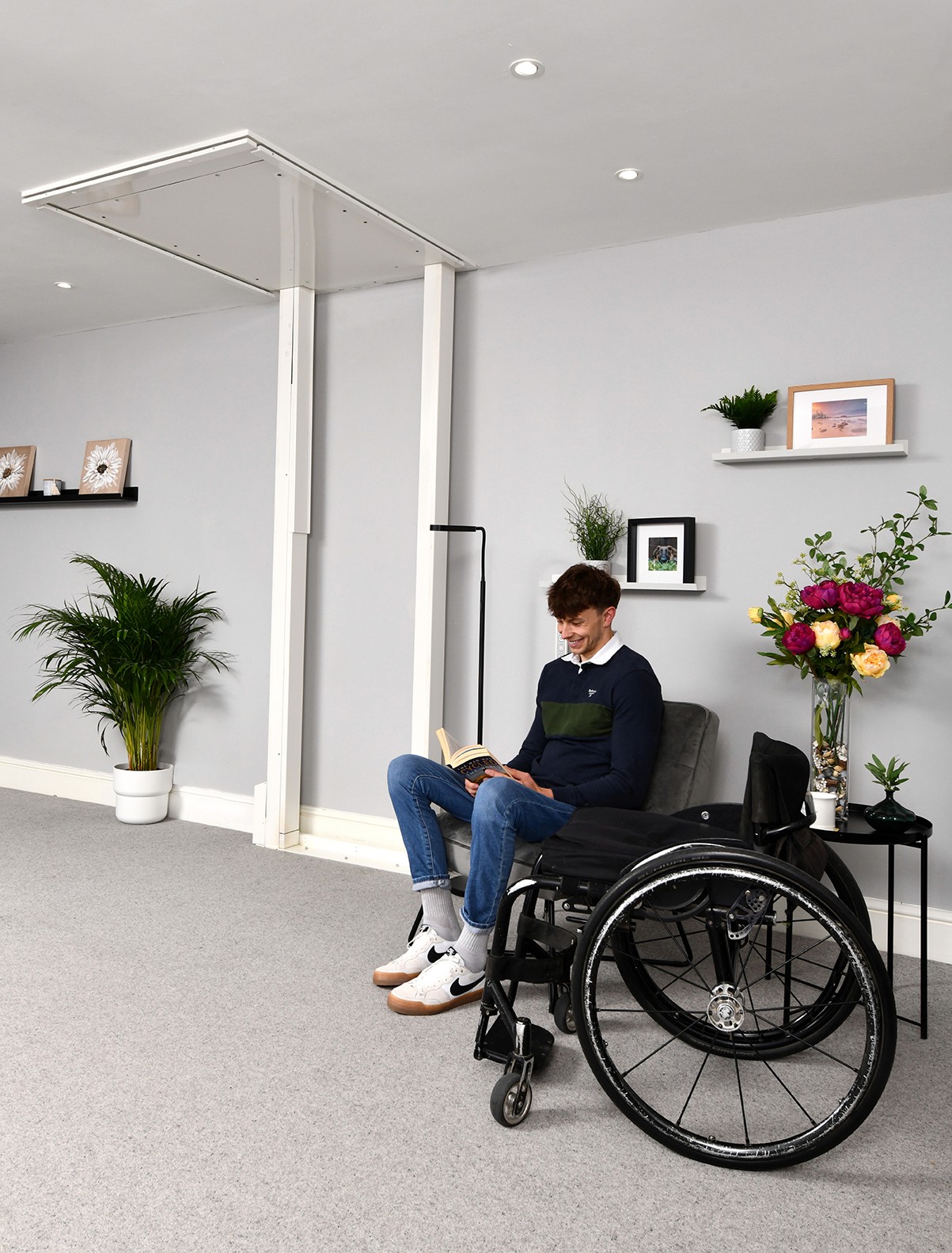 Wheelchair user seated beside an Elevate AVA Reach through-floor homelift at upper floor level in a UK residential room — white aluminium frame with ceiling aperture visible above