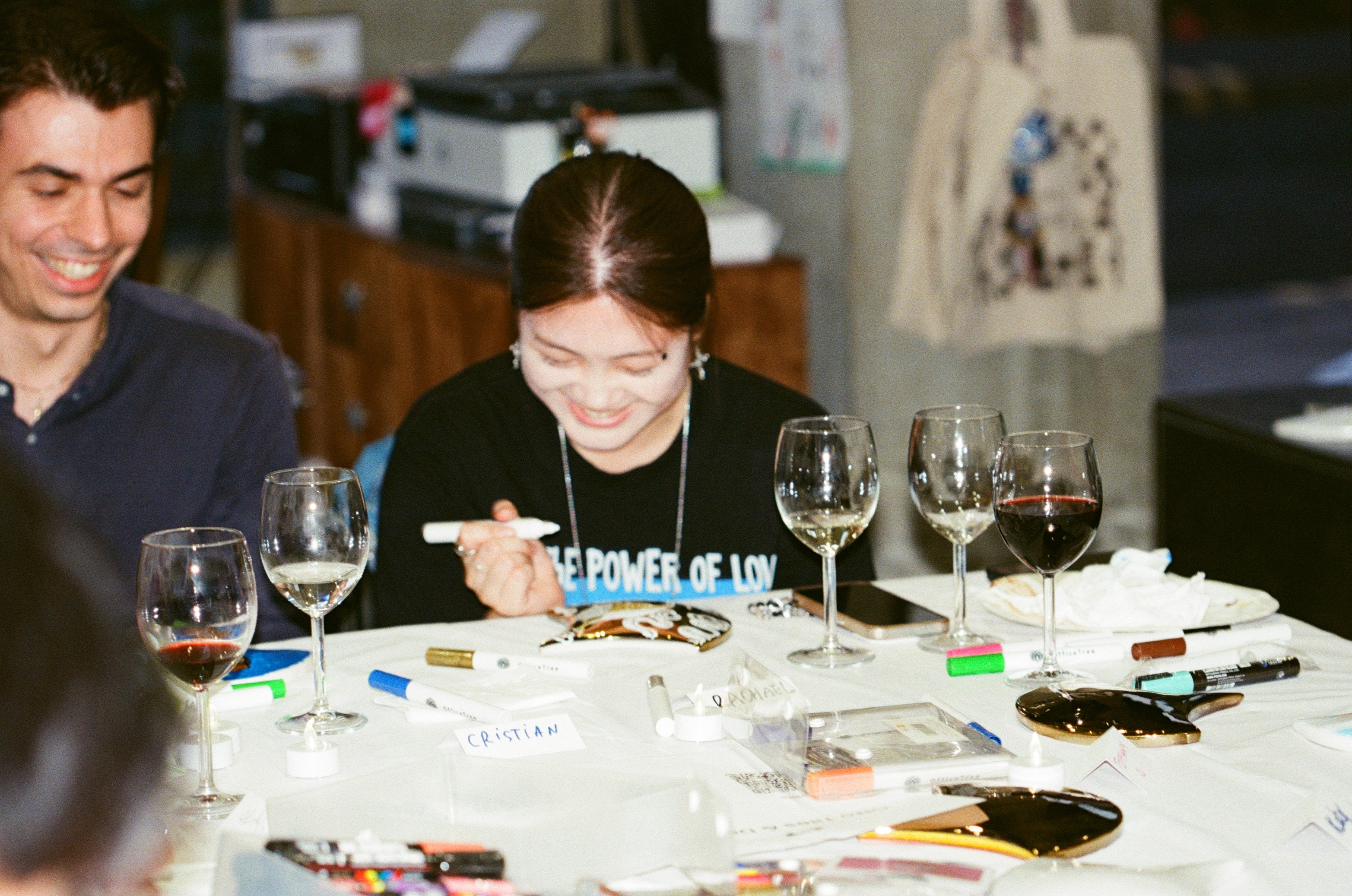 A guest smiling as she paints a gold fan tile with a white marker, another guest laughing beside her, wine glasses, scattered markers and handwritten place cards across the table.