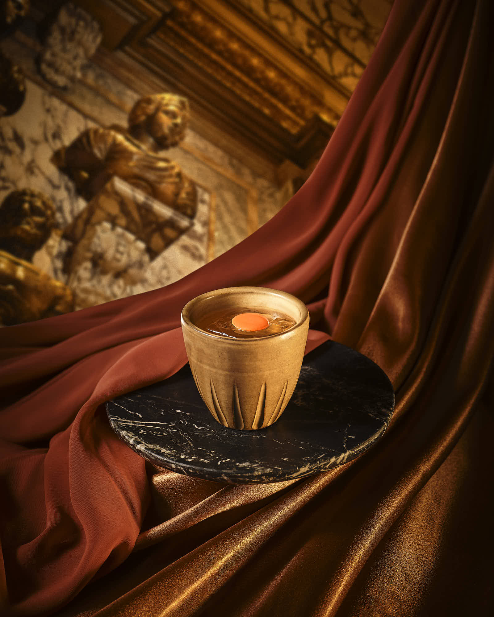 A cocktail in a ceramic cup is set against rich, draped fabrics and wooden elements, creating a cozy atmosphere.