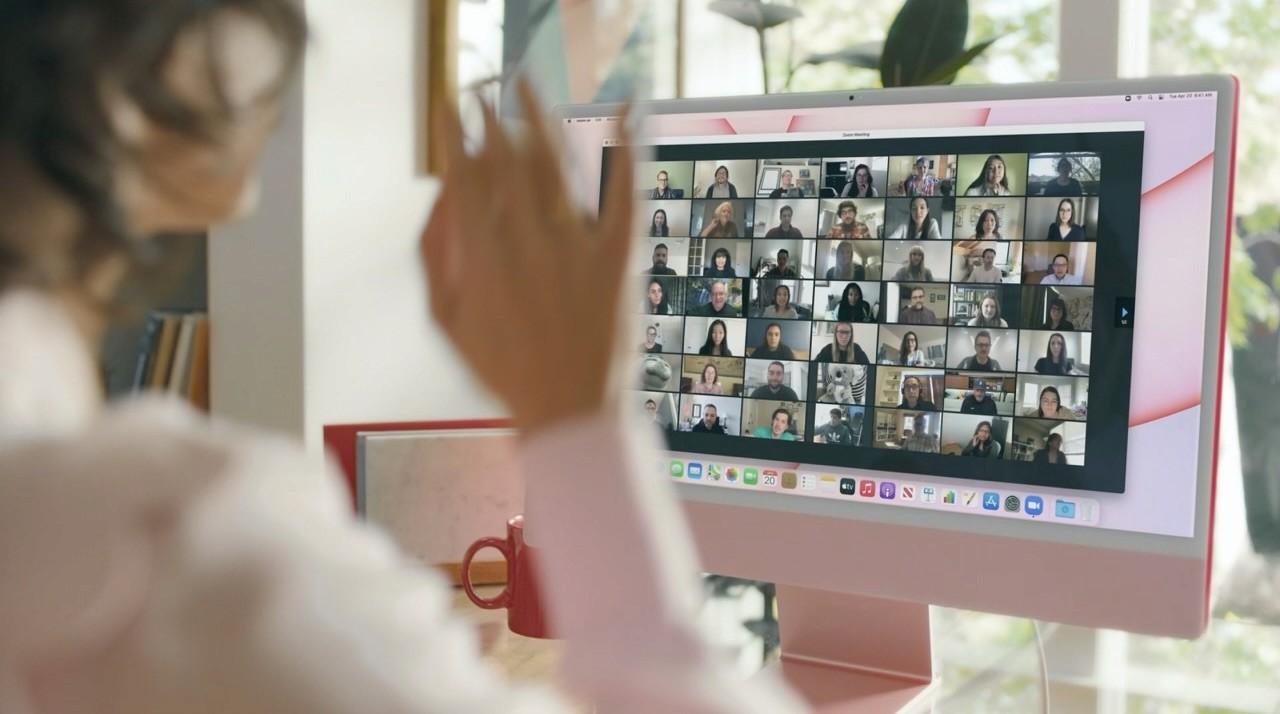 Person waving at an iMac showing a large video call grid, representing remote collaboration and team communication.