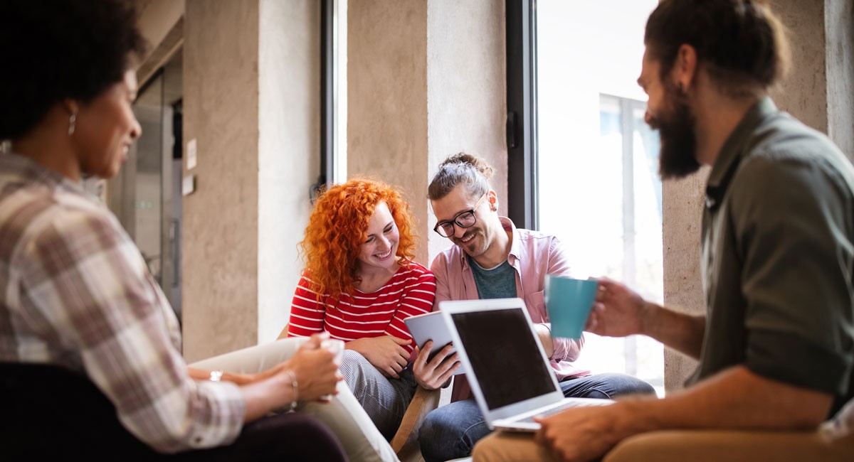 Four colleagues smiling and collaborating casually with a tablet and coffee in a modern office