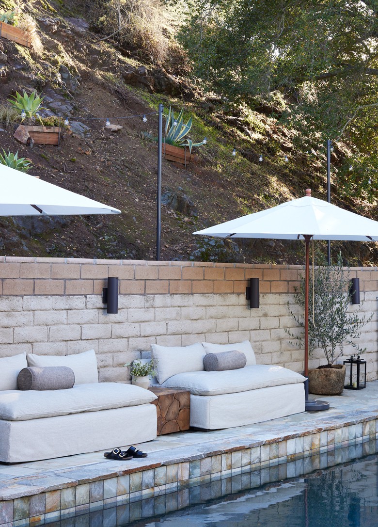 This outdoor living design blends modern luxury with nature, featuring a beach-club-style seating area, natural wood tones, lush greenery, and elegant pool stone accents for a tranquil retreat