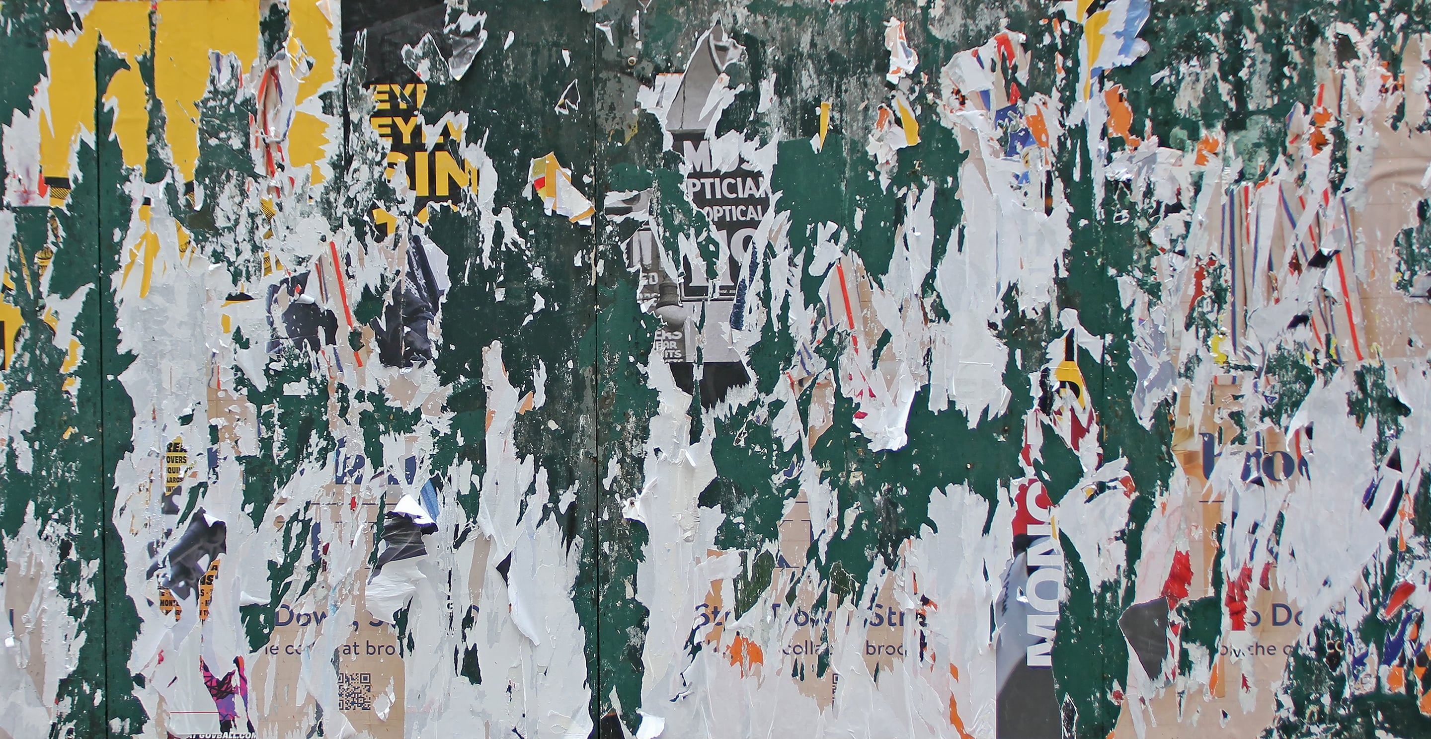 Photograph of a weathered wall covered in torn posters, with layers of green paint, paper, and printed text exposed beneath peeling surfaces. The composition reveals an abstract interplay of color, texture, and urban decay.