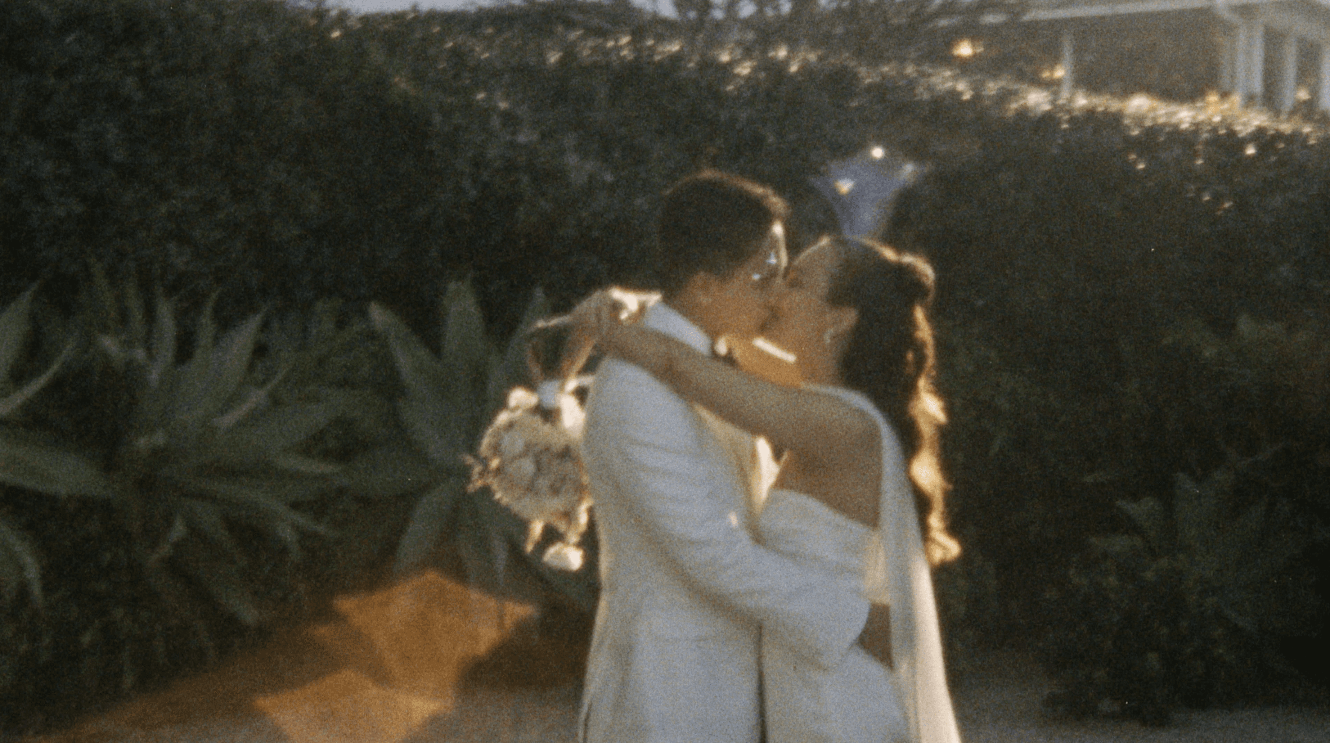 A couple stands facing each other, smiling and exchanging vows under the dappled sunlight of an outdoor garden, surrounded by greenery and seated guests in formal attire.