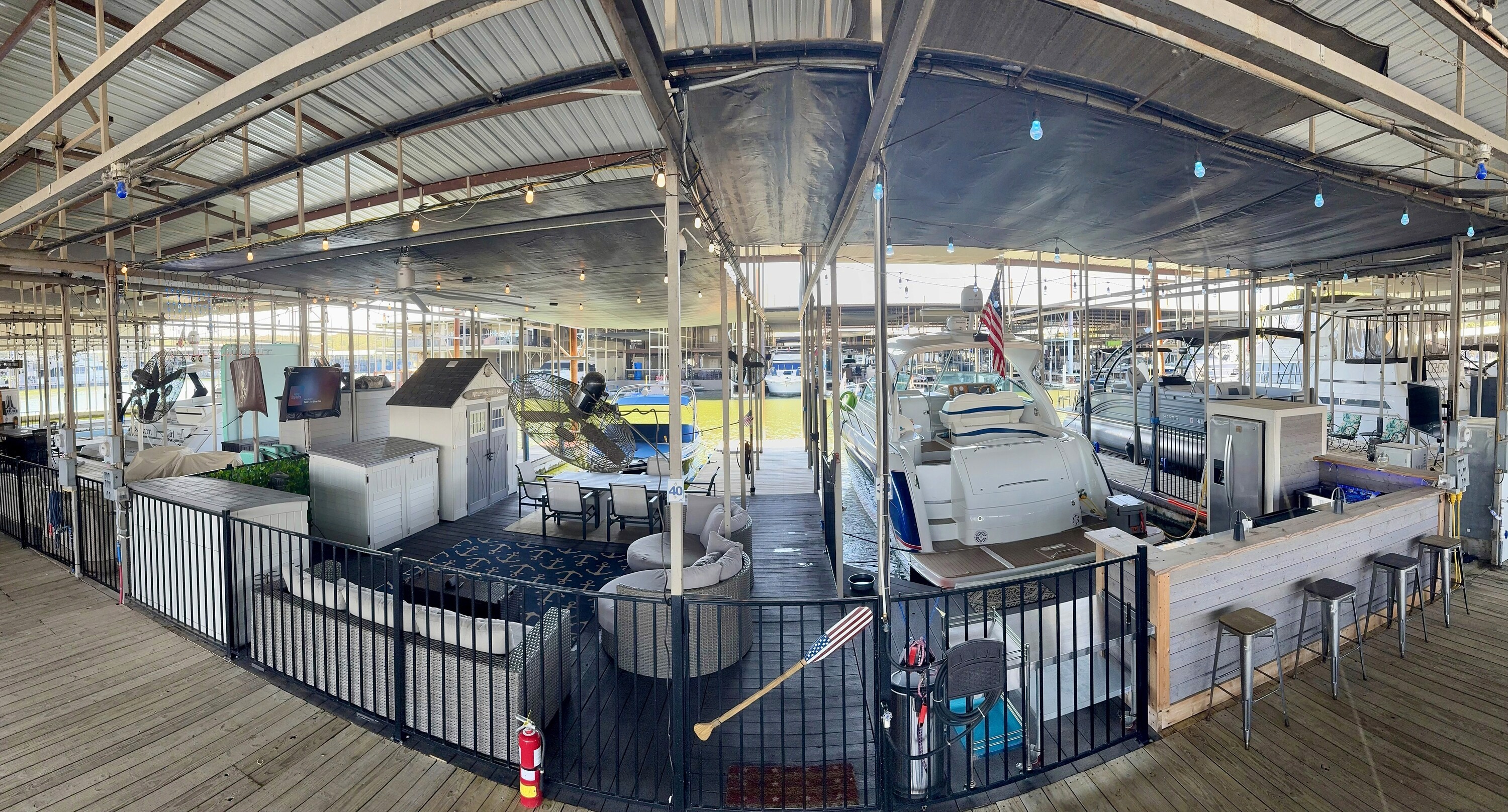 Panoramic view of a spacious marina with multiple docked boats covered by a metal roof, featuring cozy seating areas, tables, and nautical decor, including an American flag and a golf club, against a backdrop of bright lights and water.