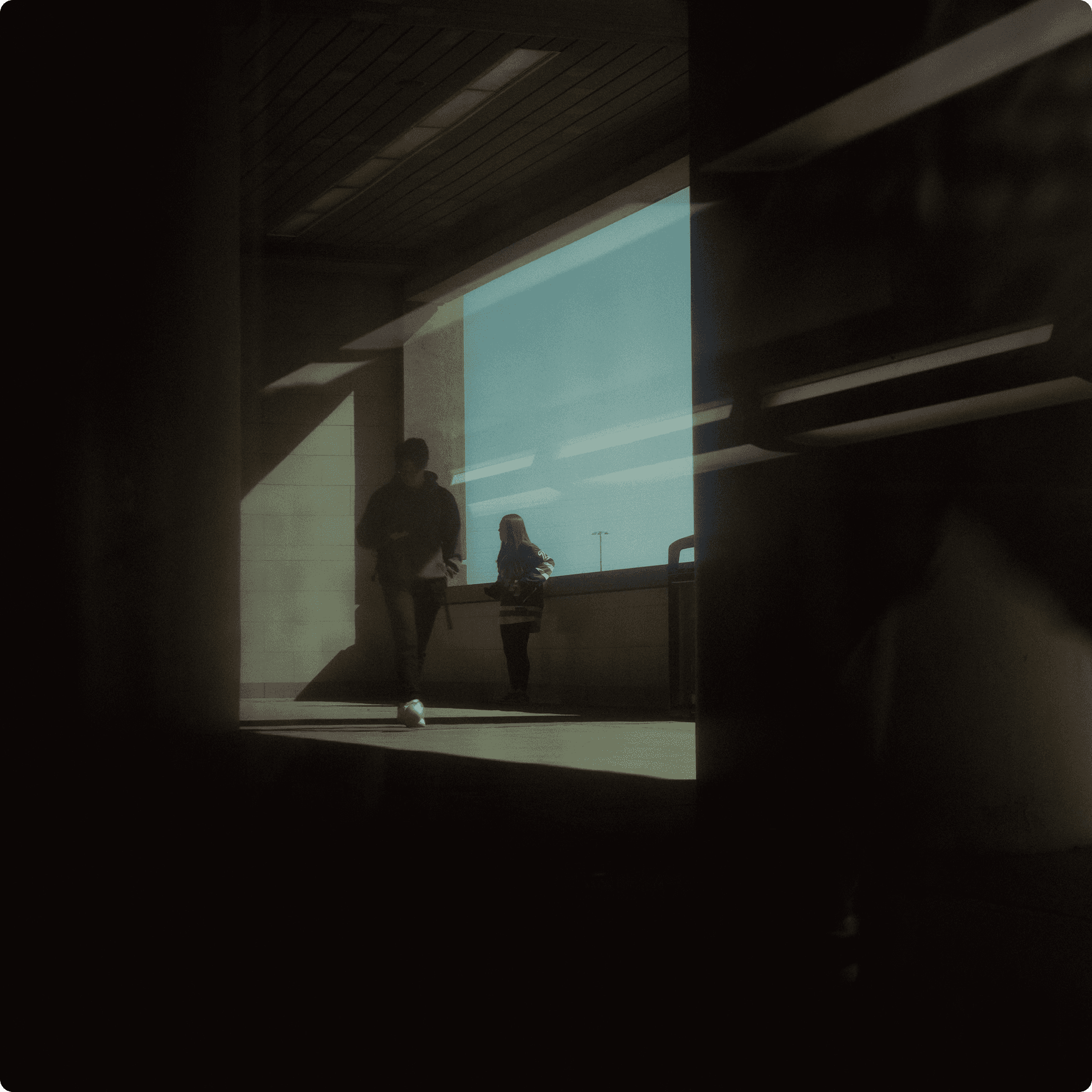 Two people walking through a dimly lit building walkway with bright natural sunlight streaming in
