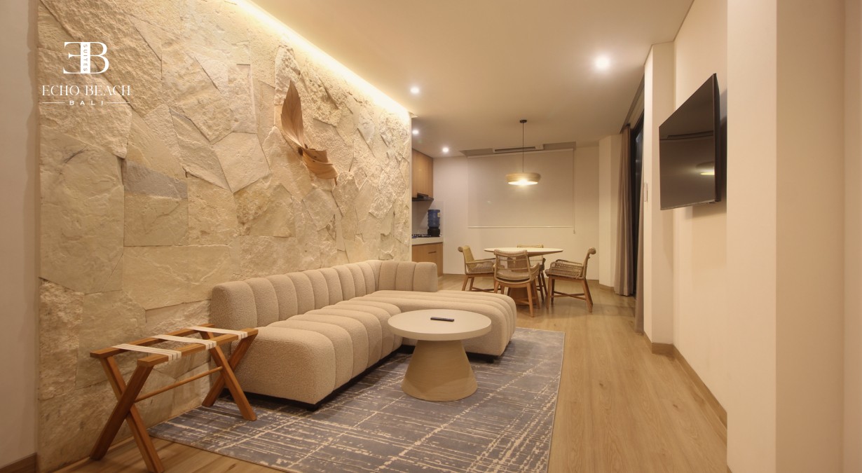 A warm, modern living room at Echo Beach Suite in Canggu, Bali, featuring a natural stone accent wall, an L-shaped beige sofa, a round coffee table, a textured rug, and a minimalist dining area in the background, reflecting the calm ambiance near Echo Beach.