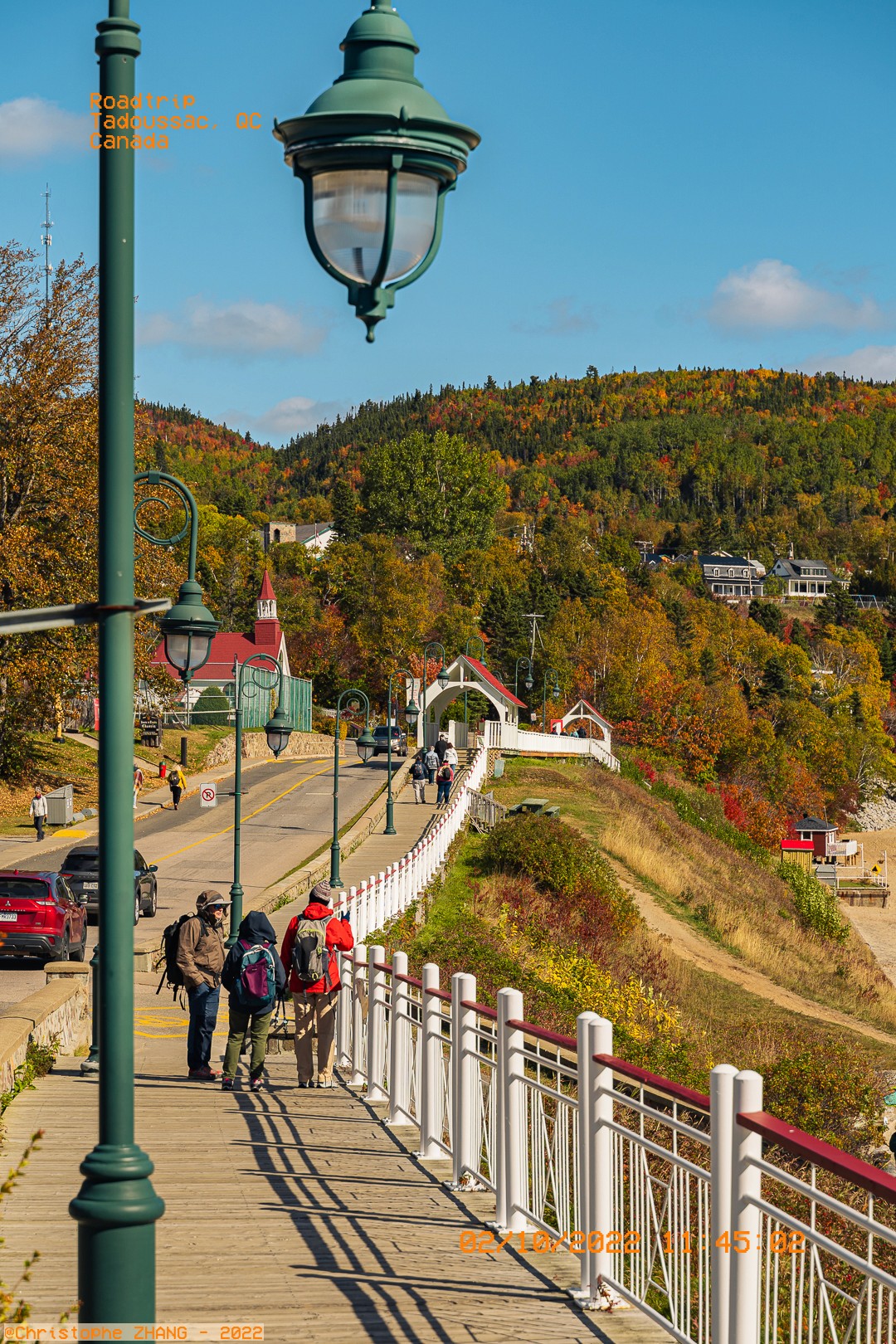 Promeneurs sur une passerelle en bois à Tadoussac avec des collines automnales en arrière-plan.