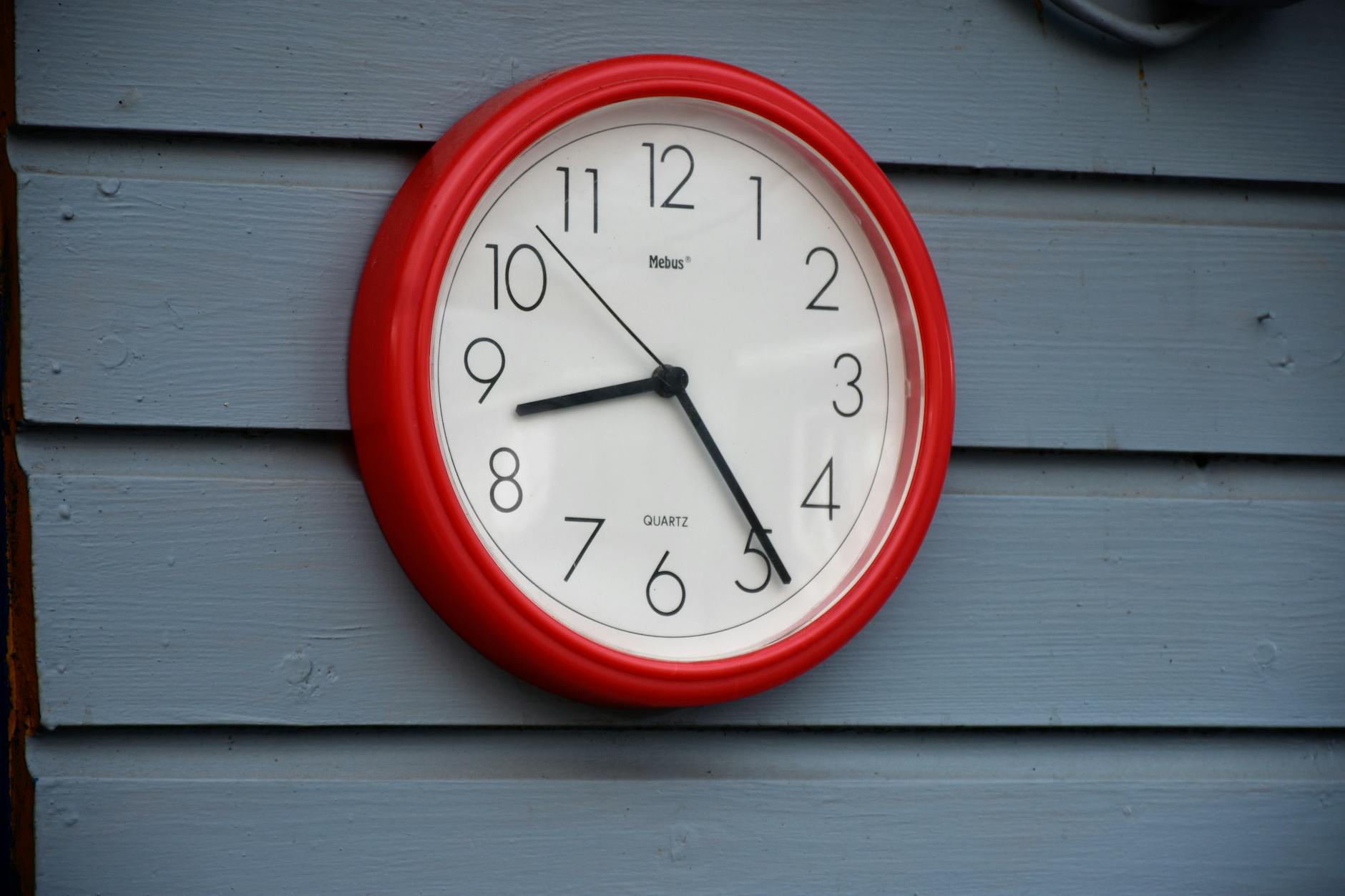 A red wall clock with black hands on a blue wooden wall, captured in daylight.