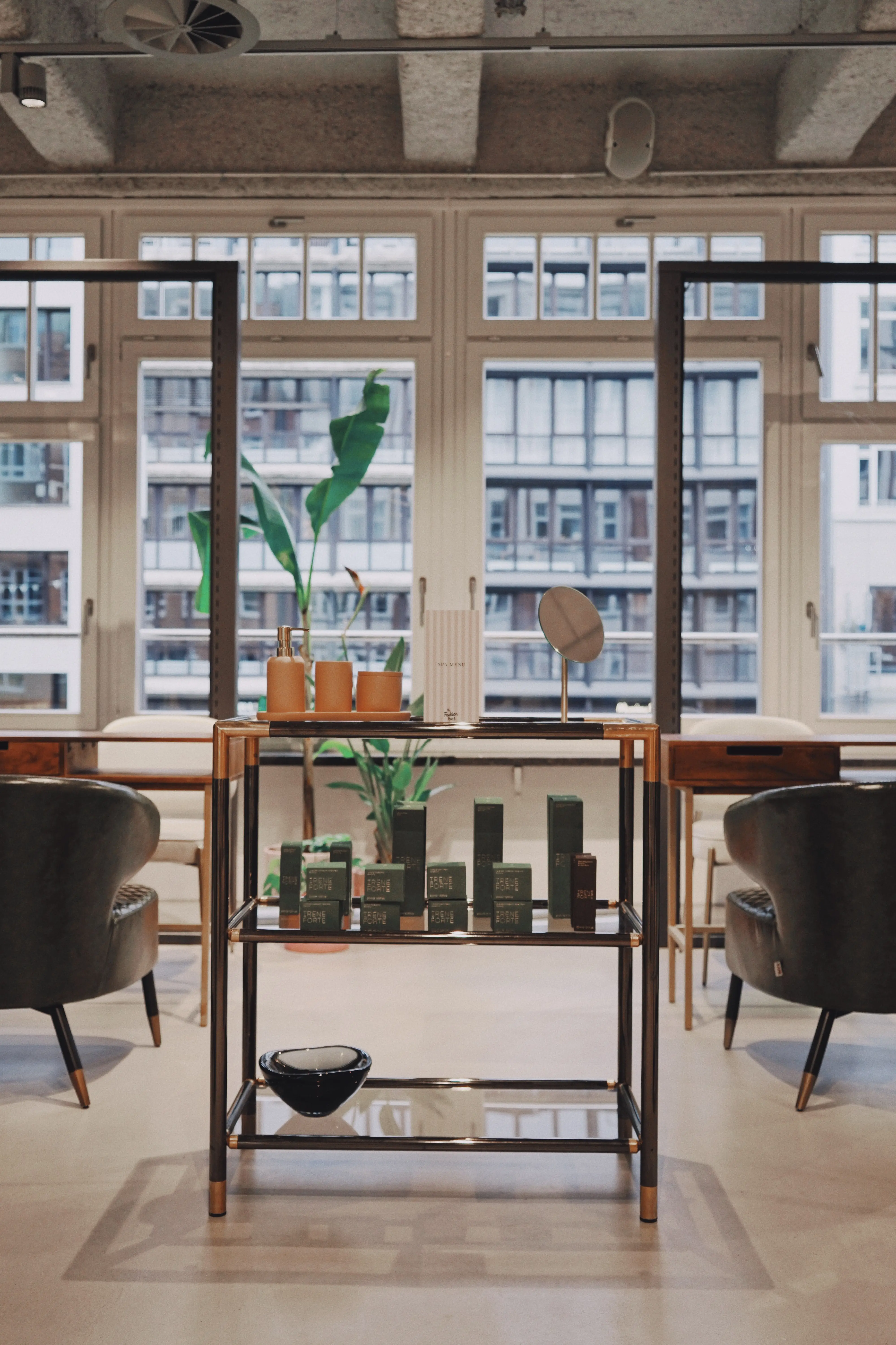 Seating area at UZWEI FASHION HOTEL with two dark green velvet armchairs flanking a chrome-and-glass shelf with green spa products, in front of tall factory windows.