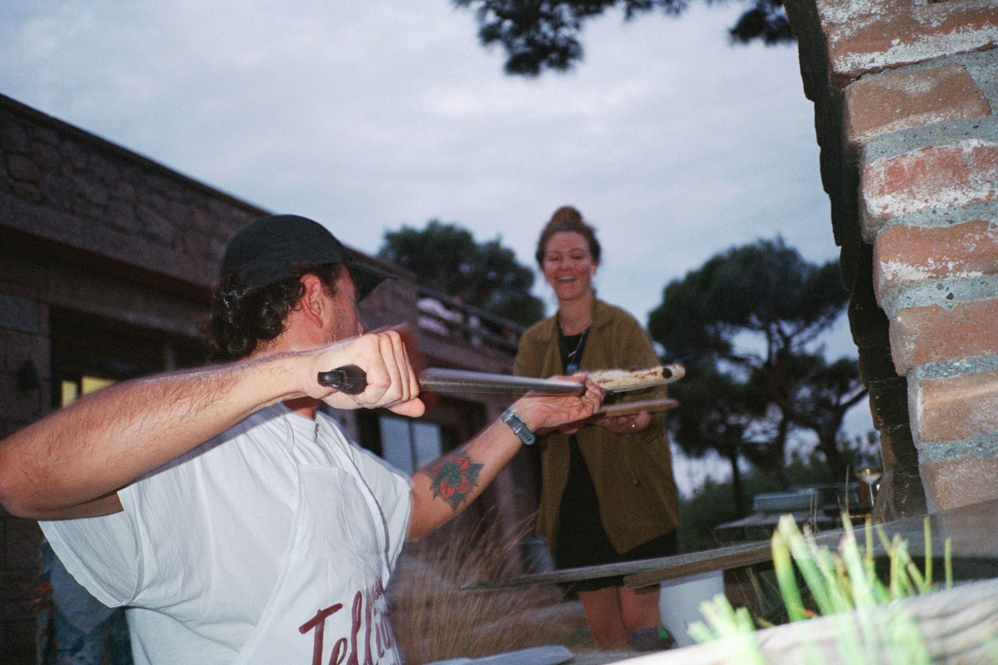 A person with tattoos, in a white shirt and black cap, is using a long-handled tool to stoke a fire in an outdoor brick fireplace, while another person, smiling, stands nearby amidst a natural setting with trees and a rustic building in the background.