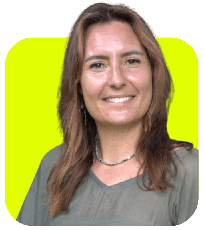 A woman with long, curly hair sits by a large window, smiling and wearing a black top and blue jeans.