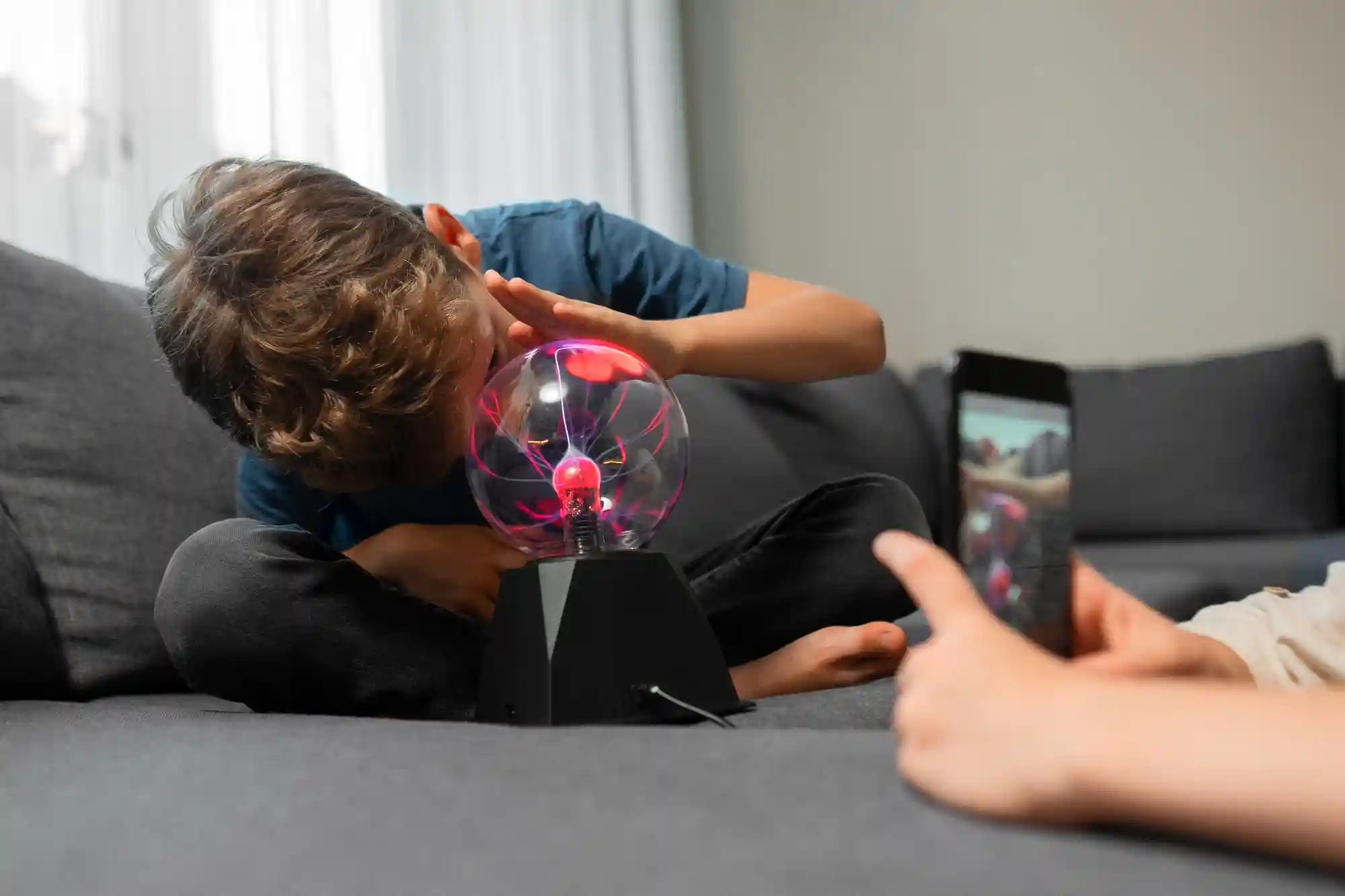 A child observes a glowing plasma ball lamp while another person captures the scientific moment on a smartphone.