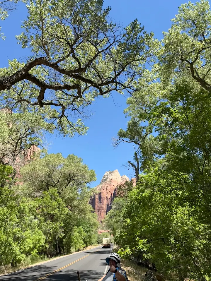 Sara smiling and giving a thumbs up while riding an electric bike, with trees overhead and mountains in the background on the road to The Narrows.