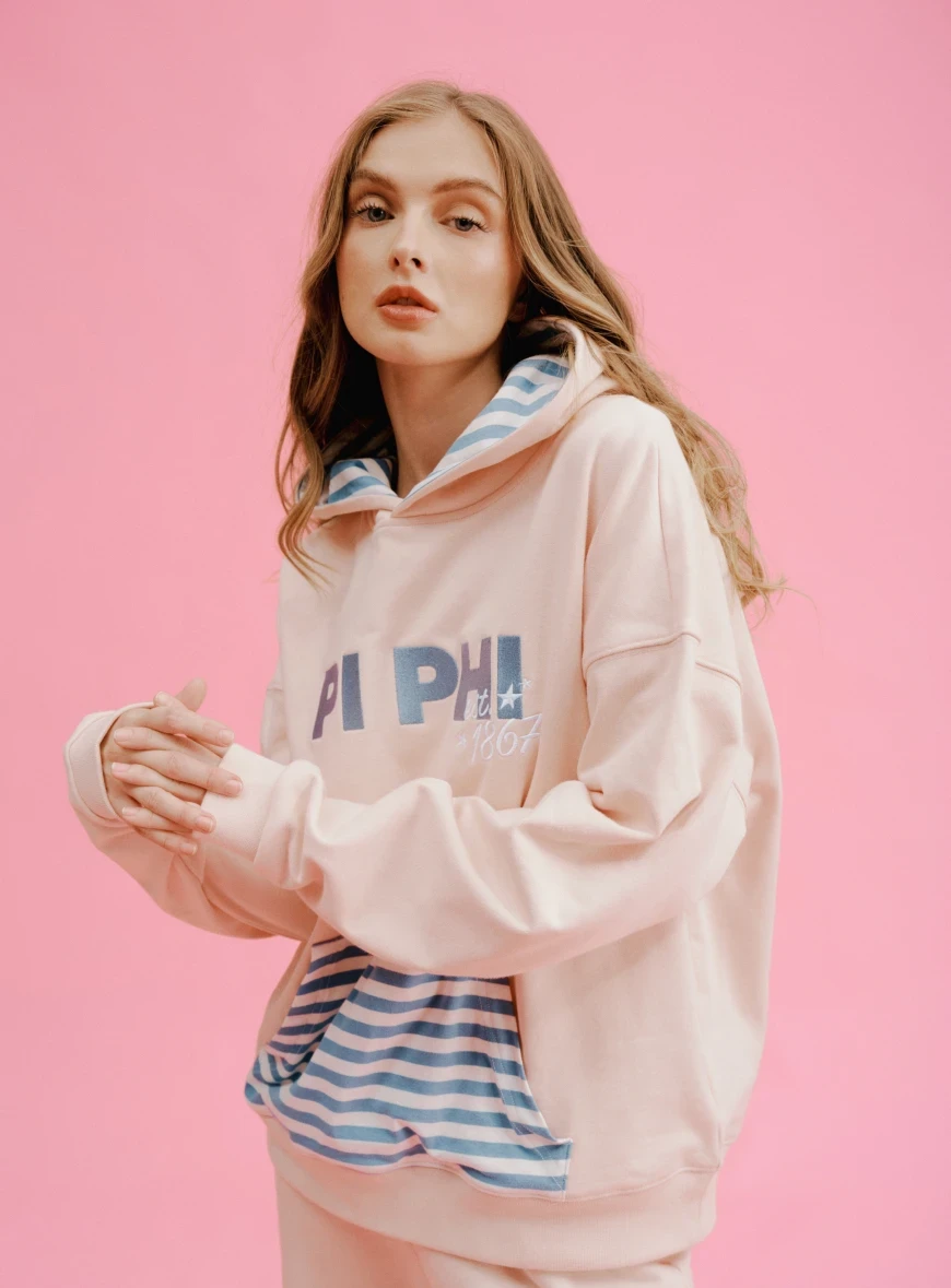 Model wearing a pale pink sorority hoodie with a blue-and-white striped front pocket and inner hood.