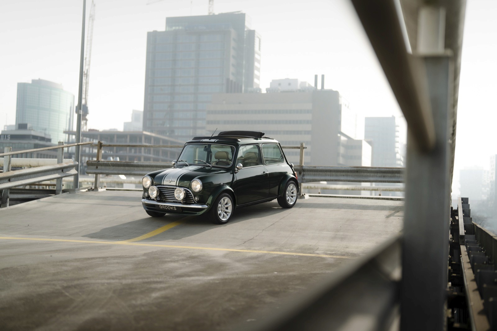 A dark green mini classic car parked on an elevated road, with city buildings in the background under a hazy sky.