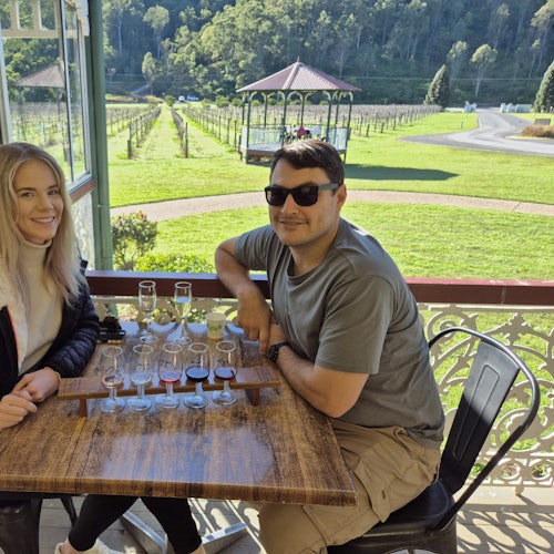 Een gestreepte luchtballon zweeft over een grasvlakte bij zonsondergang. In de verte is nog een andere ballon zichtbaar in de lucht.