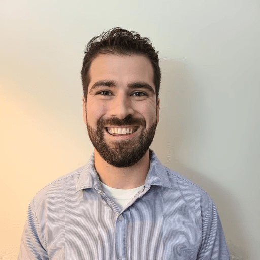 A person with curly hair and glasses, wearing an orange sweater, looks thoughtfully into the distance, resting their chin on their hand. Warm, cozy ambiance