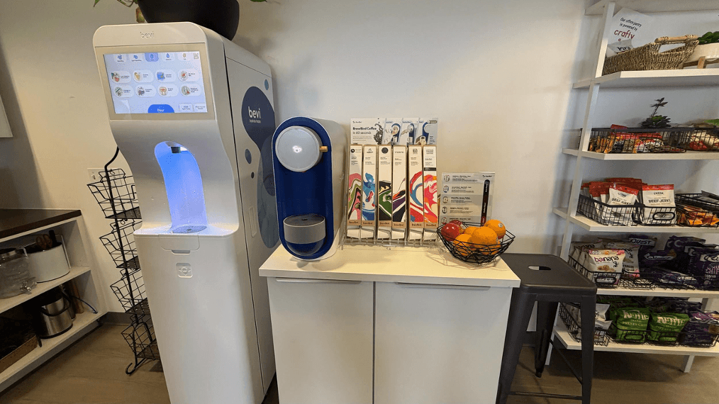 Title: BrewBird on a standalone cabinet - Description: A blue BrewBird brewer on a compact cabinet beside a water dispenser.