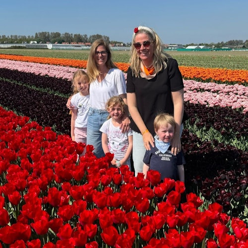 Dalawang babae at tatlong bata ang nakapose sa harap ng makukulay na hanay ng mga tulip sa isang bukid sa ilalim ng maliwanag na asul na langit.
