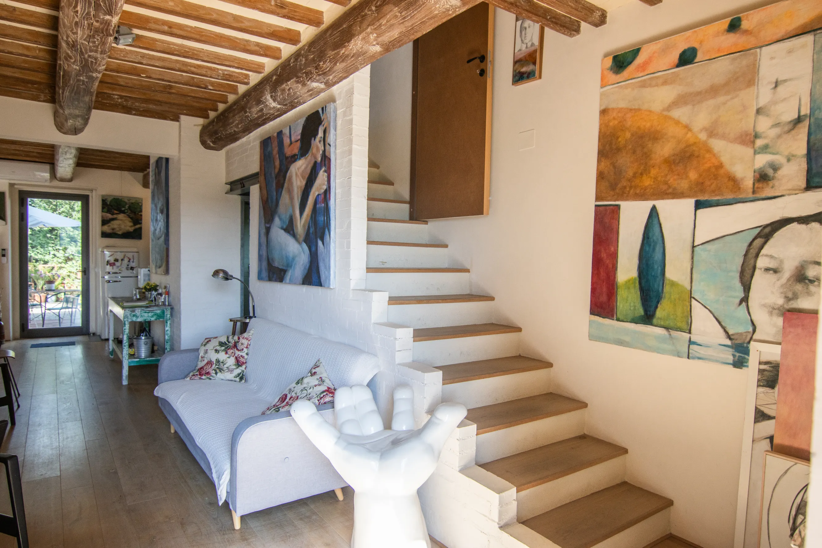 A cozy living room with wooden ceiling beams, a white sofa adorned with floral pillows, artistic paintings on the walls, and a unique hand-shaped sculpture near a staircase leading upward.