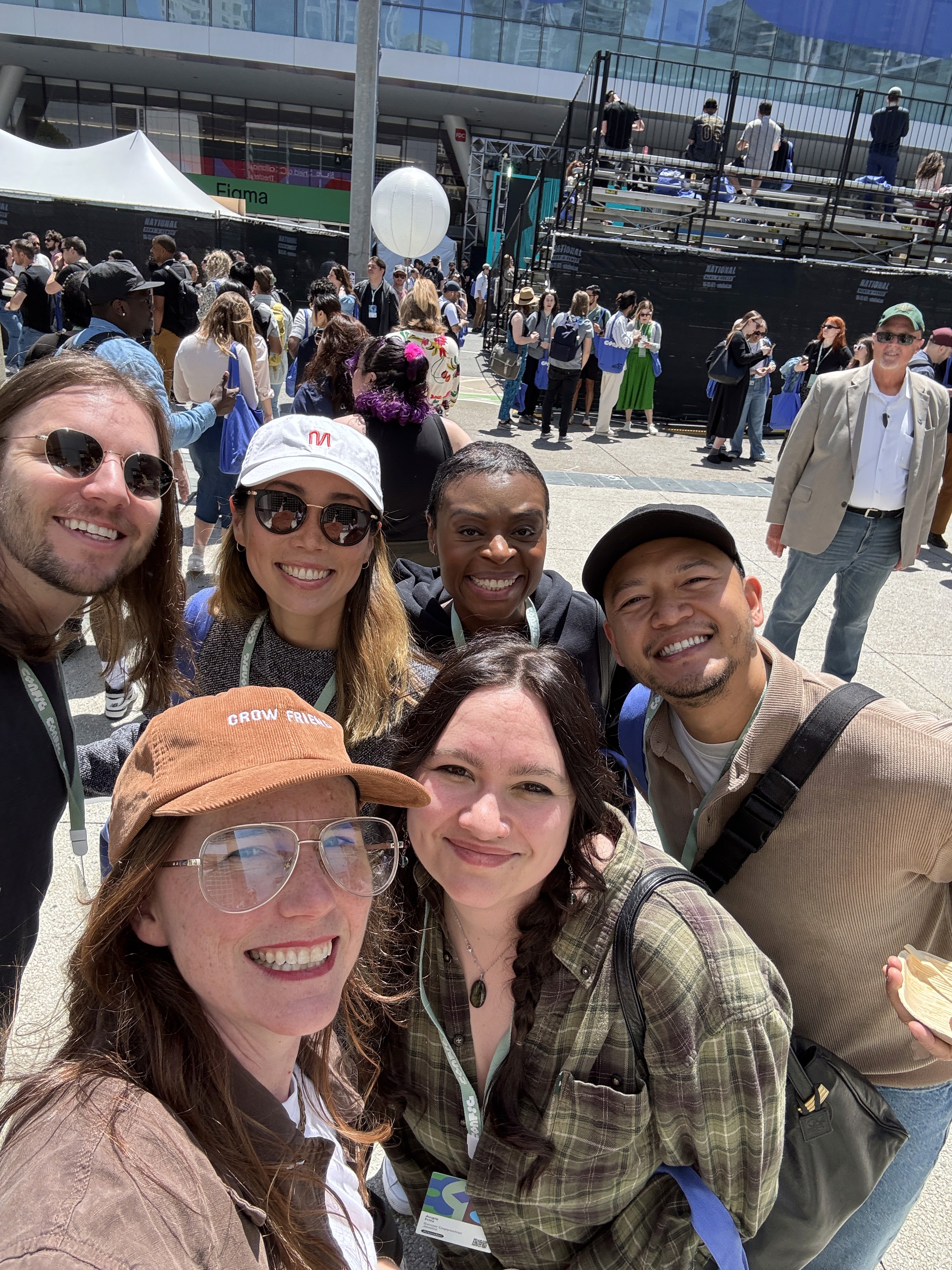 Group of smiling people taking a selfie outdoors at a crowded event, with a stage and attendees visible in the background.