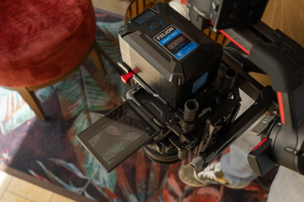 Een professionele cameraopstelling op een stabilisator legt beelden vast binnenshuis, met de focus op het display en het bedieningspaneel van de apparatuur, tegen een achtergrond met een kleurrijke stoel en een intricaat patroon tapijt.