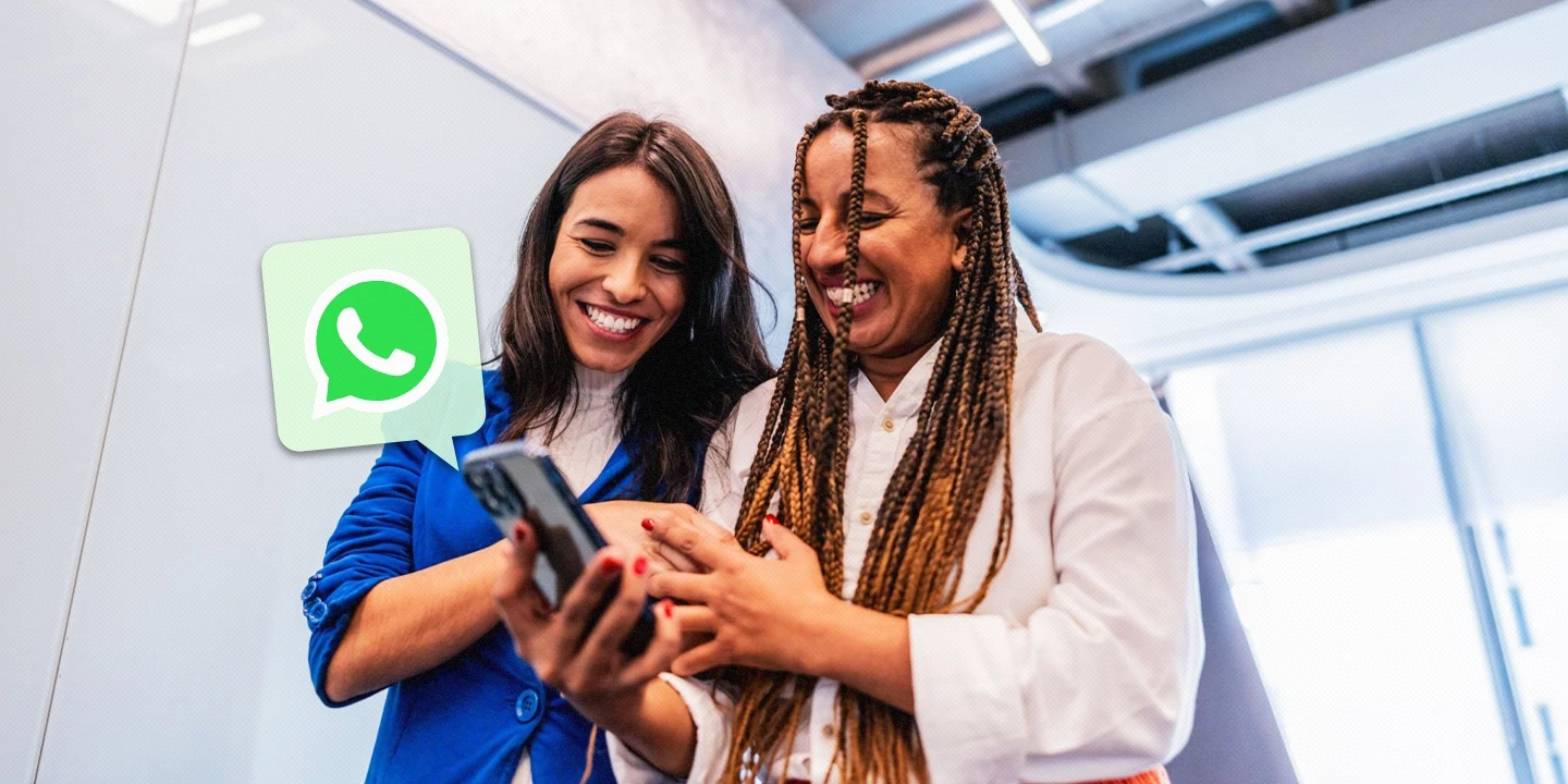 Duas mulheres sorridentes, vestindo roupas corporativas, compartilham algo em um smartphone. Um ícone do WhatsApp flutua ao lado delas, simbolizando o uso do aplicativo na comunicação empresarial