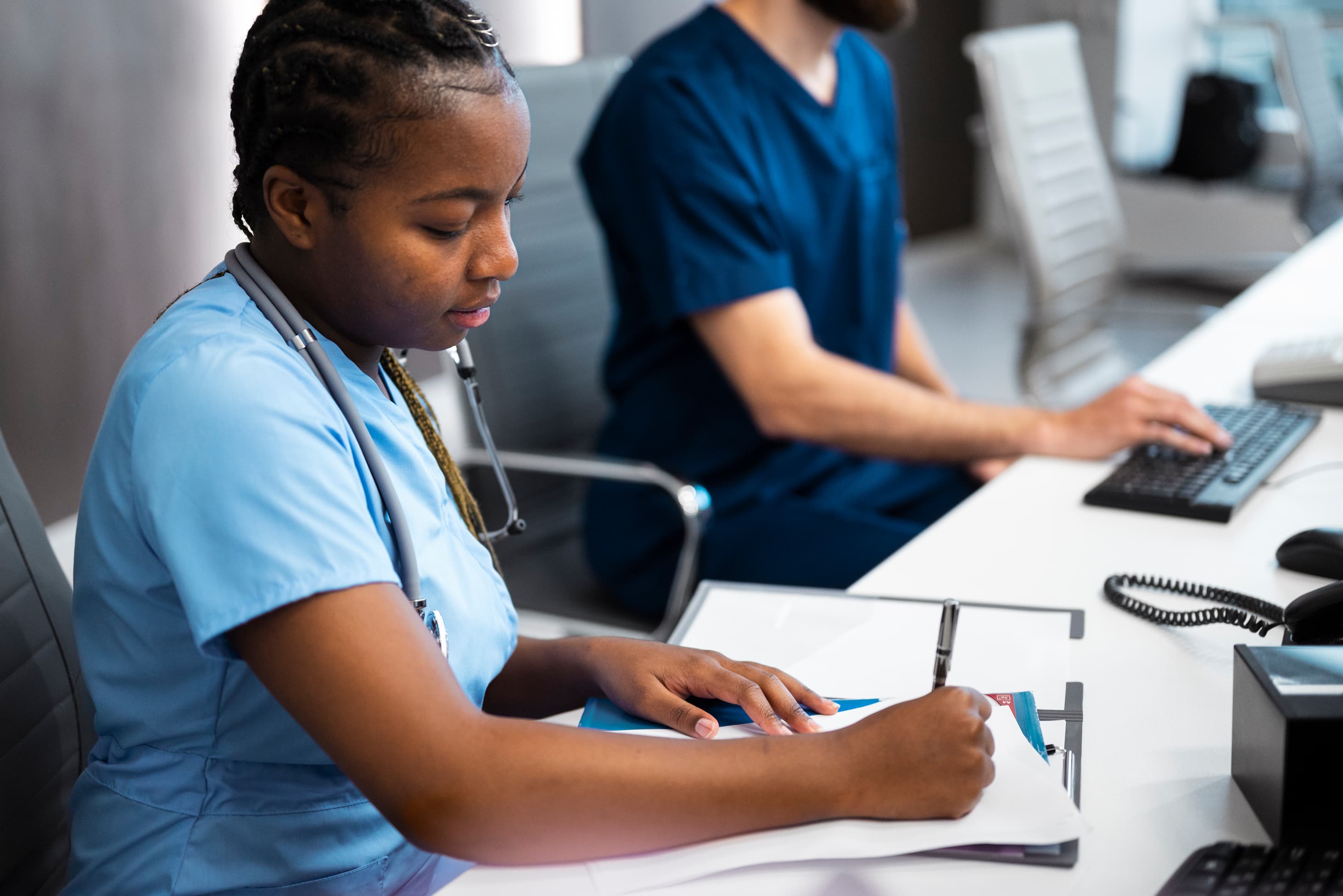 A healthcare professional in scrubs writing on a clipboard while seated at a desk, with a colleague working on a computer in the background.