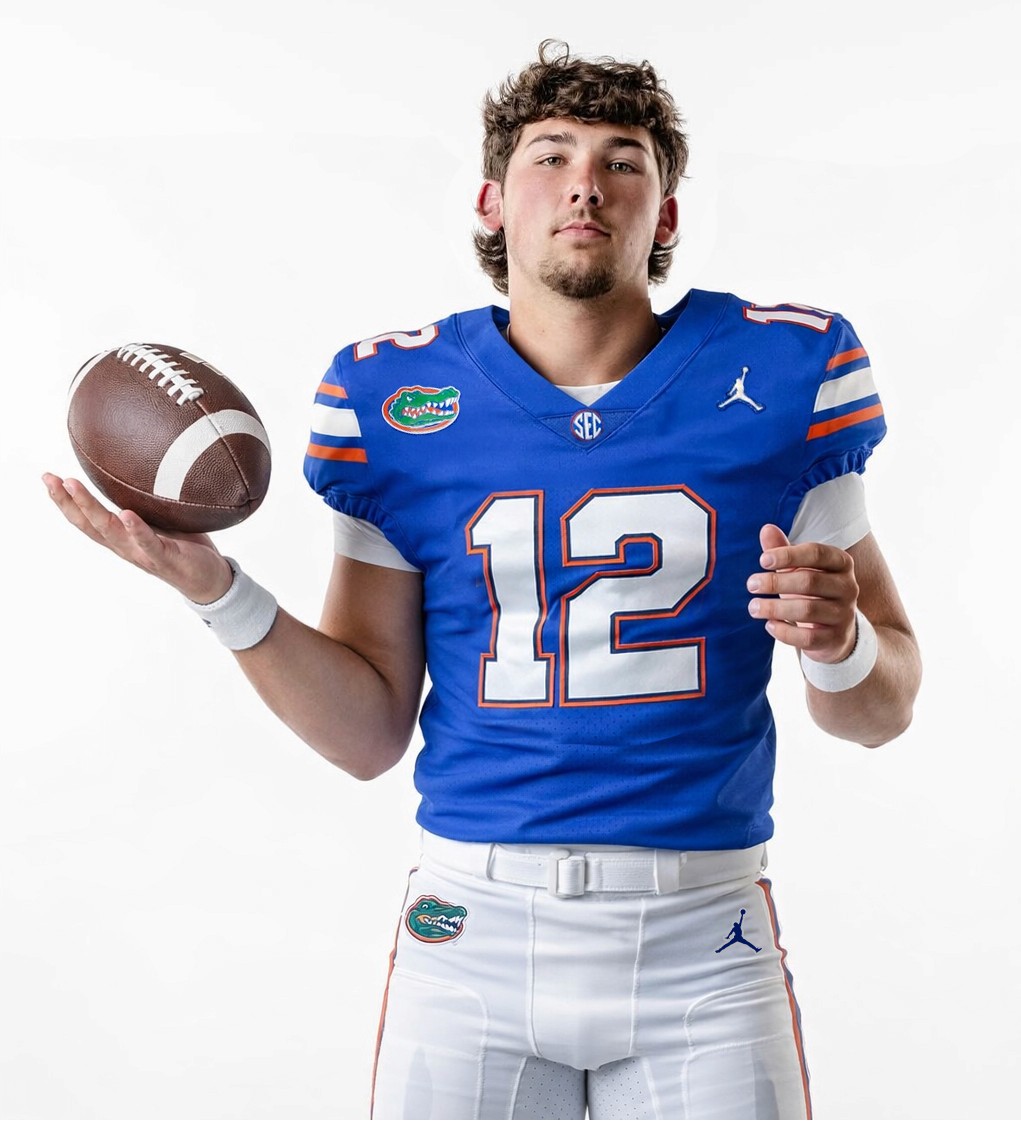 A football player in a blue Florida Gators uniform with the number 12 poses against a white background, holding a football in one hand and looking at the camera.