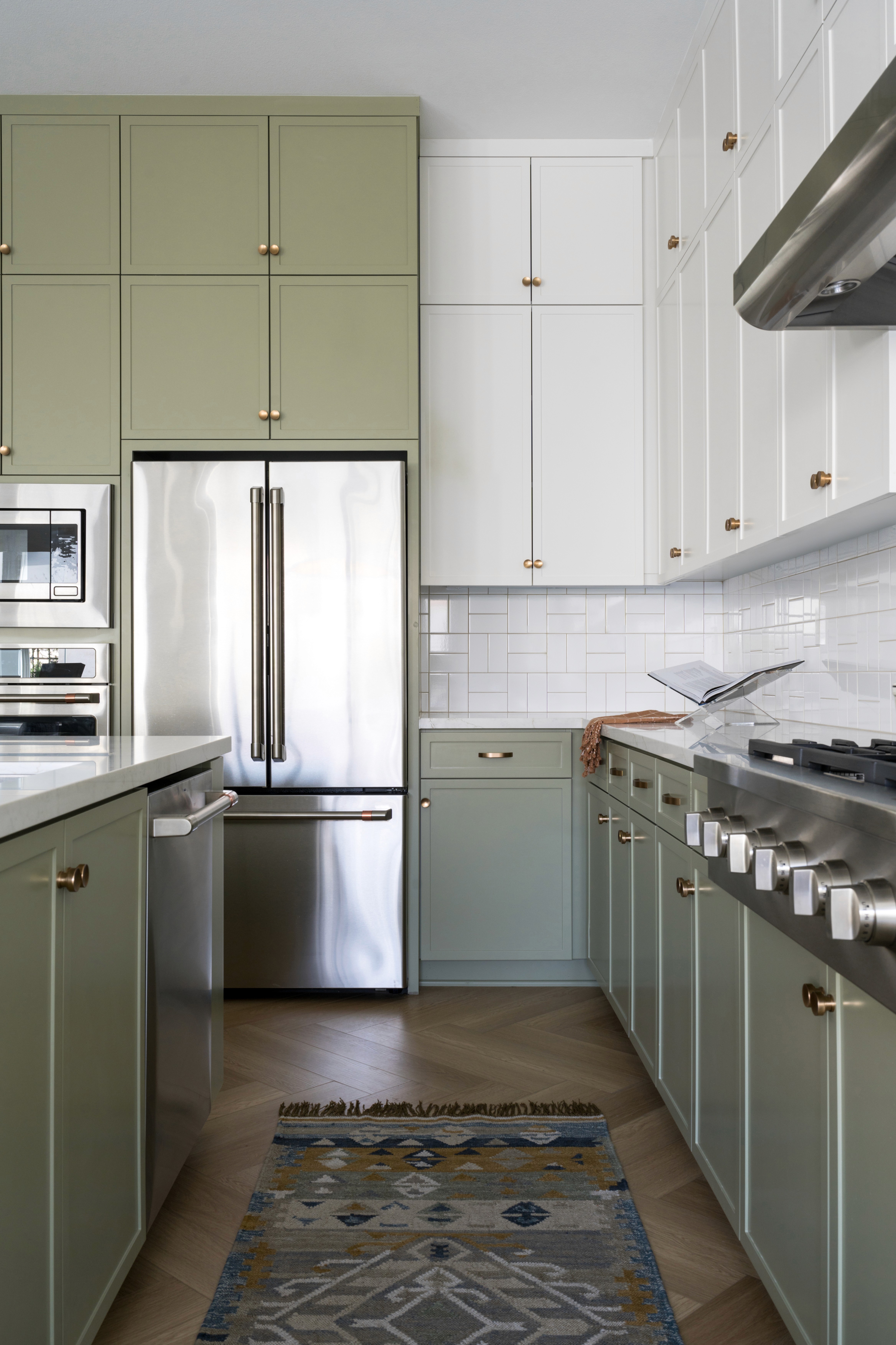 Kitchen with sage green and white cabinetry with chrome finishes, white tile backsplash, stainless steal appliances, chevron wood floor, and woven runner with shades of blue by Jubilee Interiors in Arcadia, California