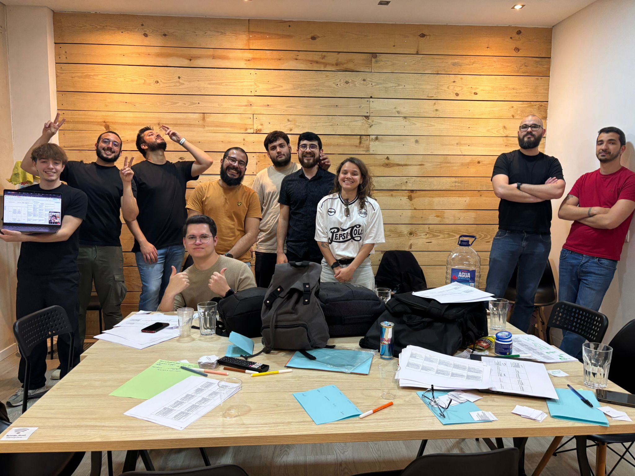 Team photo of nine men and two women in the office, while one of the man holds a computer showing his colleague remotely on the screen.