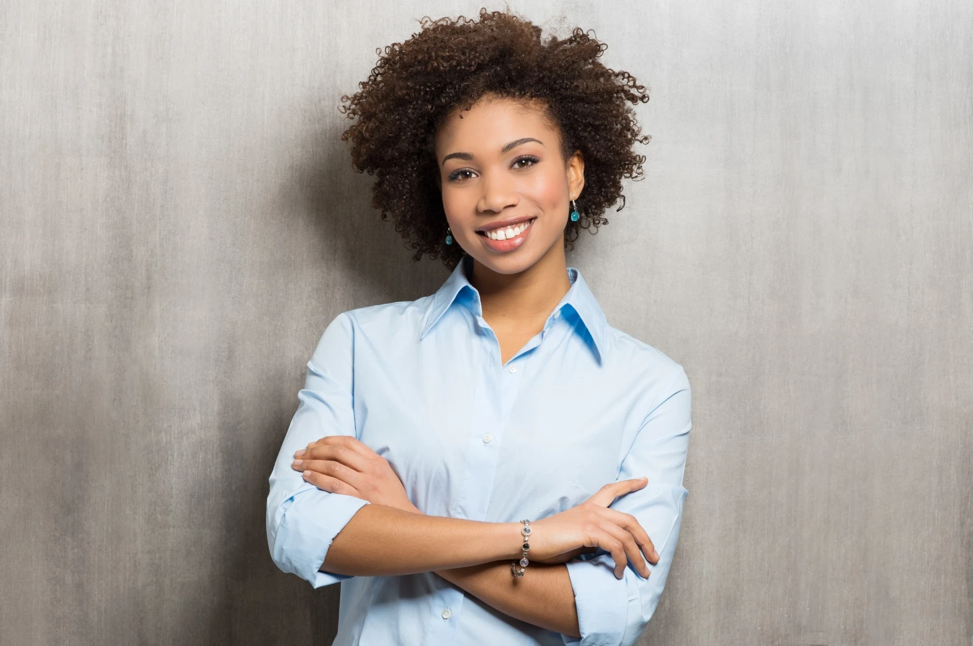 Imagem de uma mulher sorridente, com cabelos cacheados e volumosos, usando uma camisa social azul claro. Ela está de braços cruzados e possui uma expressão confiante e amigável. Ao fundo, um cenário neutro com uma parede de textura cinza, destacando a presença profissional e acolhedora dela.