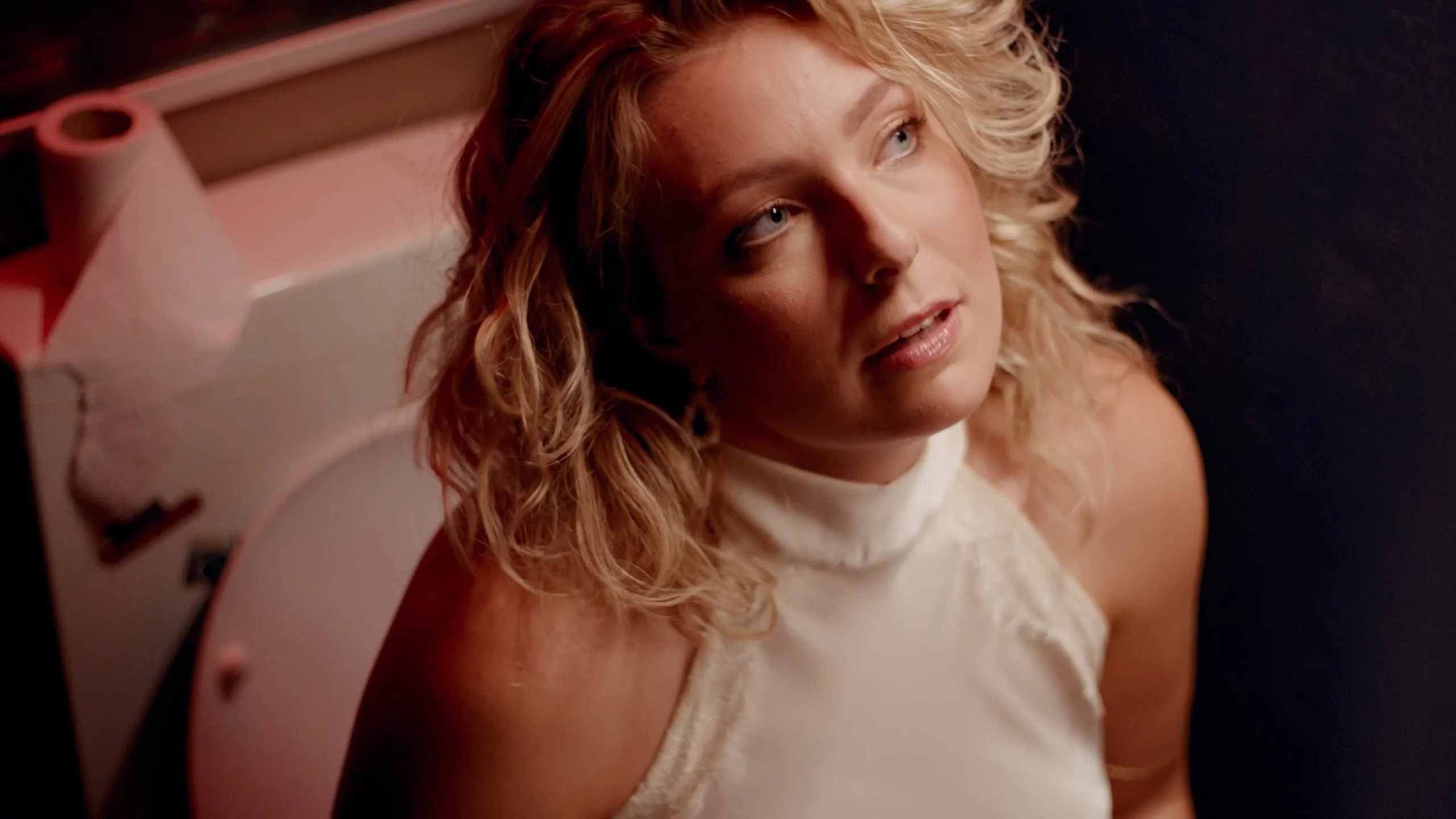 Close up portrait of a young woman with curly blonde hair, caught in warm bathroom light.