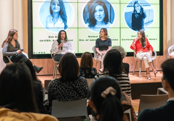 A panel of women speaking on a stage while a crowd watches