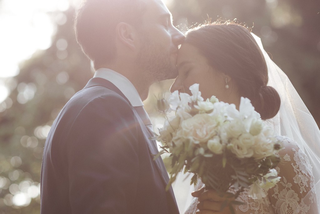 Sposi si baciano al tramonto, lei in abito bianco con bouquet di rose avorio, lui in giacca grigia, luce dorata naturale
