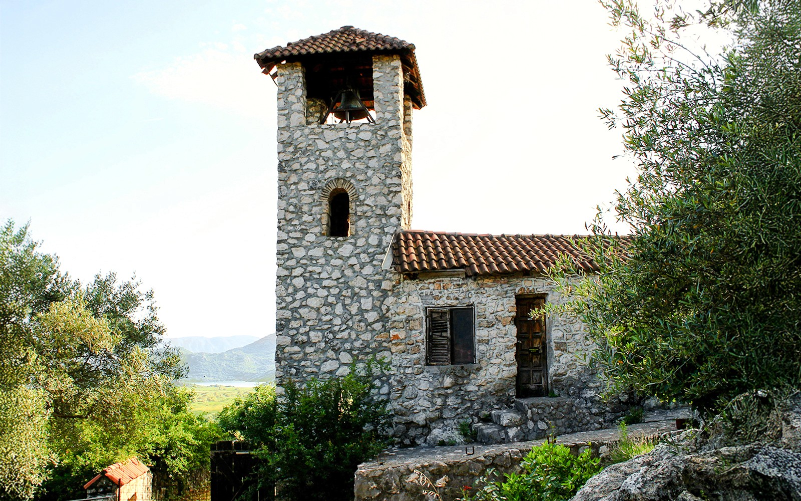 Каменная колокольня монастыря Ком на озере Скадар, окружённая зеленью.