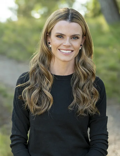 A smiling young woman in a pink blouse stands in an office setting, with soft lighting and a professional backdrop.