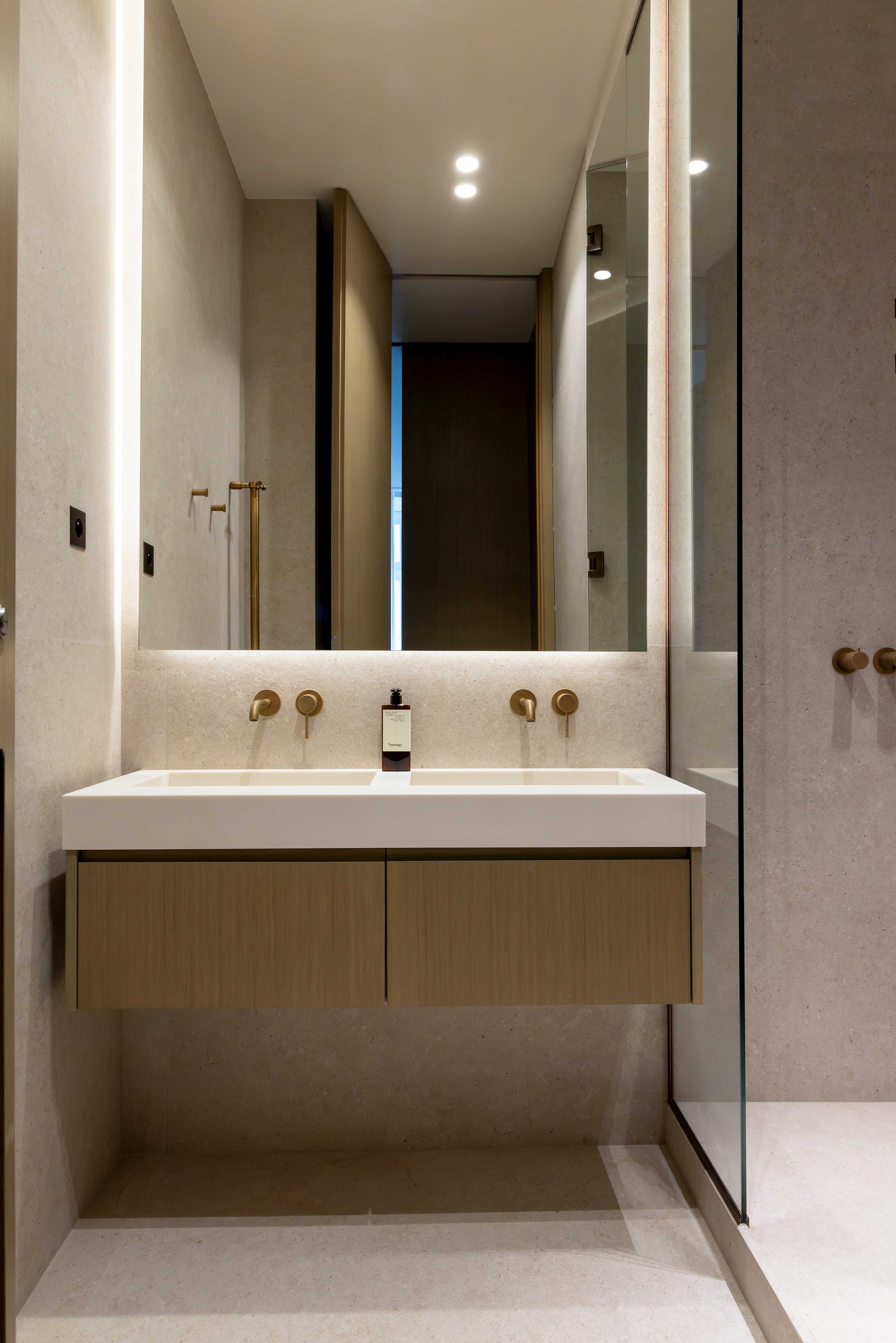 Salle de bain avec double vasque suspendue, robinetterie doree, grand miroir retroeclaire et paroi vitree