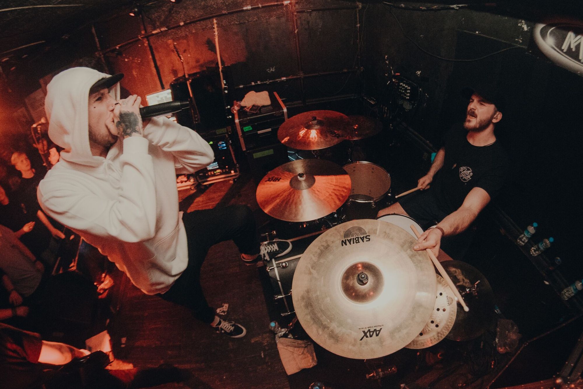 Singer in a white hoodie and drummer performing in a small venue