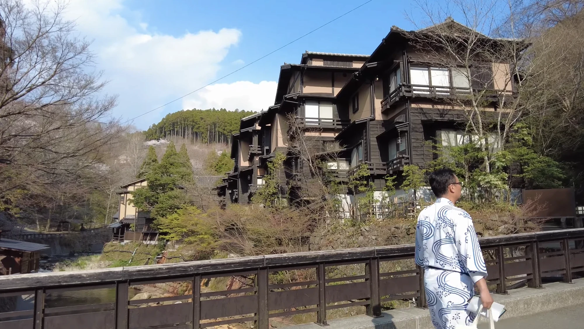 A guest in a blue and white patterned yukata pausing on the bridge at Kurokawa Onsen, inns rising behind