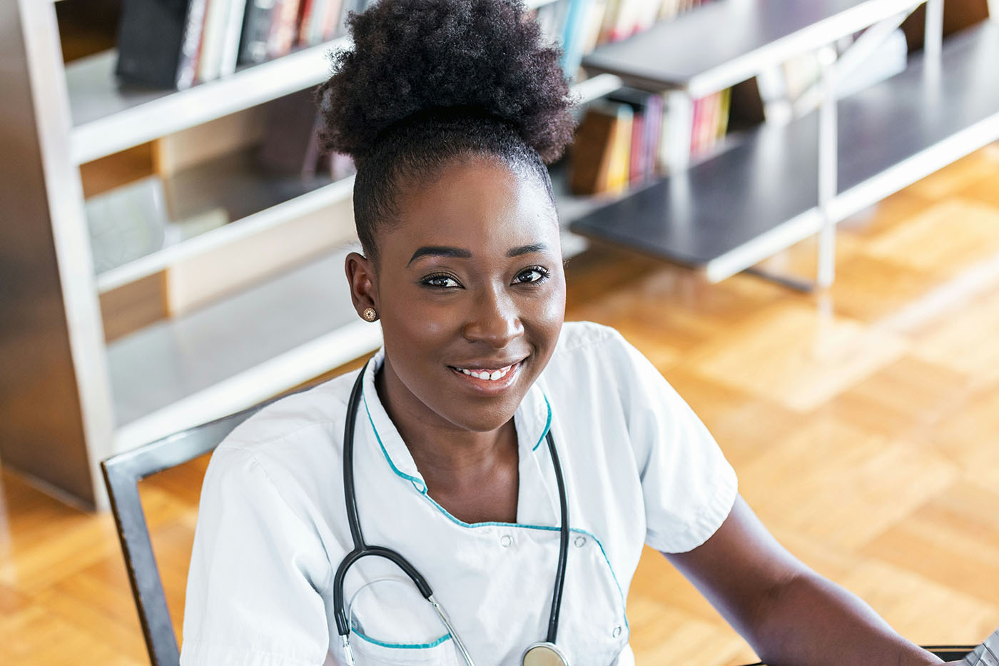 A smiling Black woman wearing a white medical uniform and stethoscope sitting in a library.