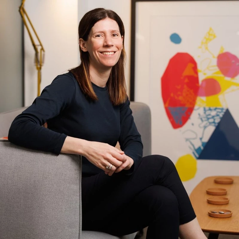 A person sits on a grey sofa in a modern living space wearing a dark long‑sleeved top and black trousers. A large abstract artwork and a small wooden table add colour and texture to the room.