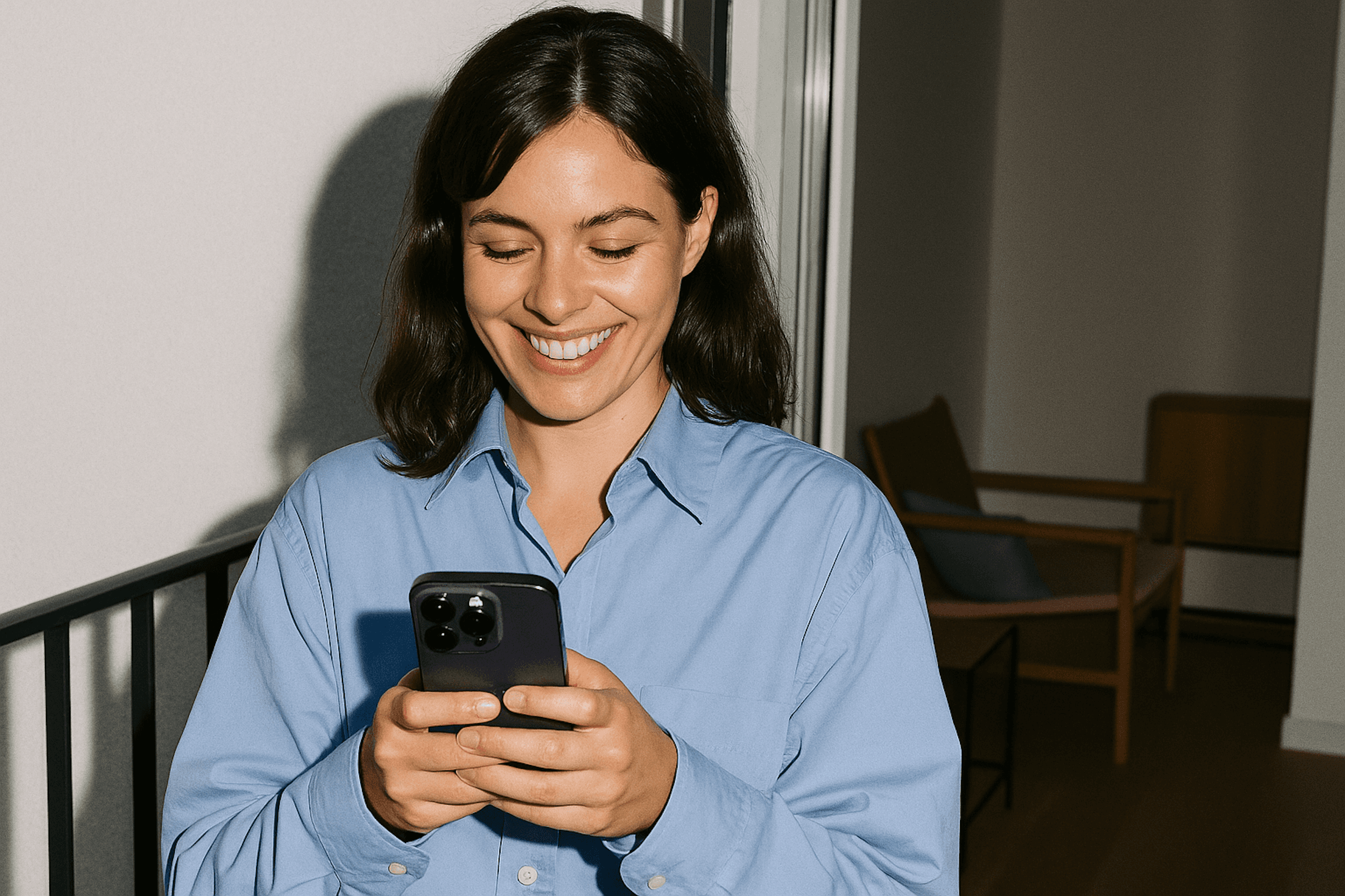 Smiling woman in a light blue shirt using a smartphone indoors, with a modern and cozy background setting.