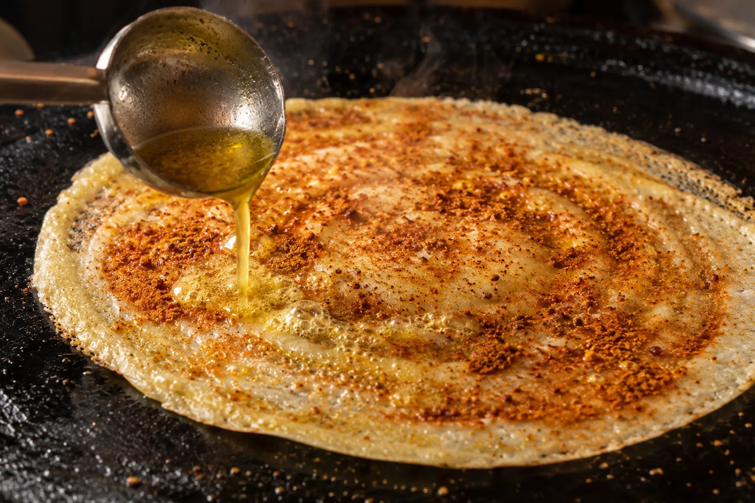 Hot ghee poured from a ladle onto a podi dosa cooking on a tawa at Jun Restaurant