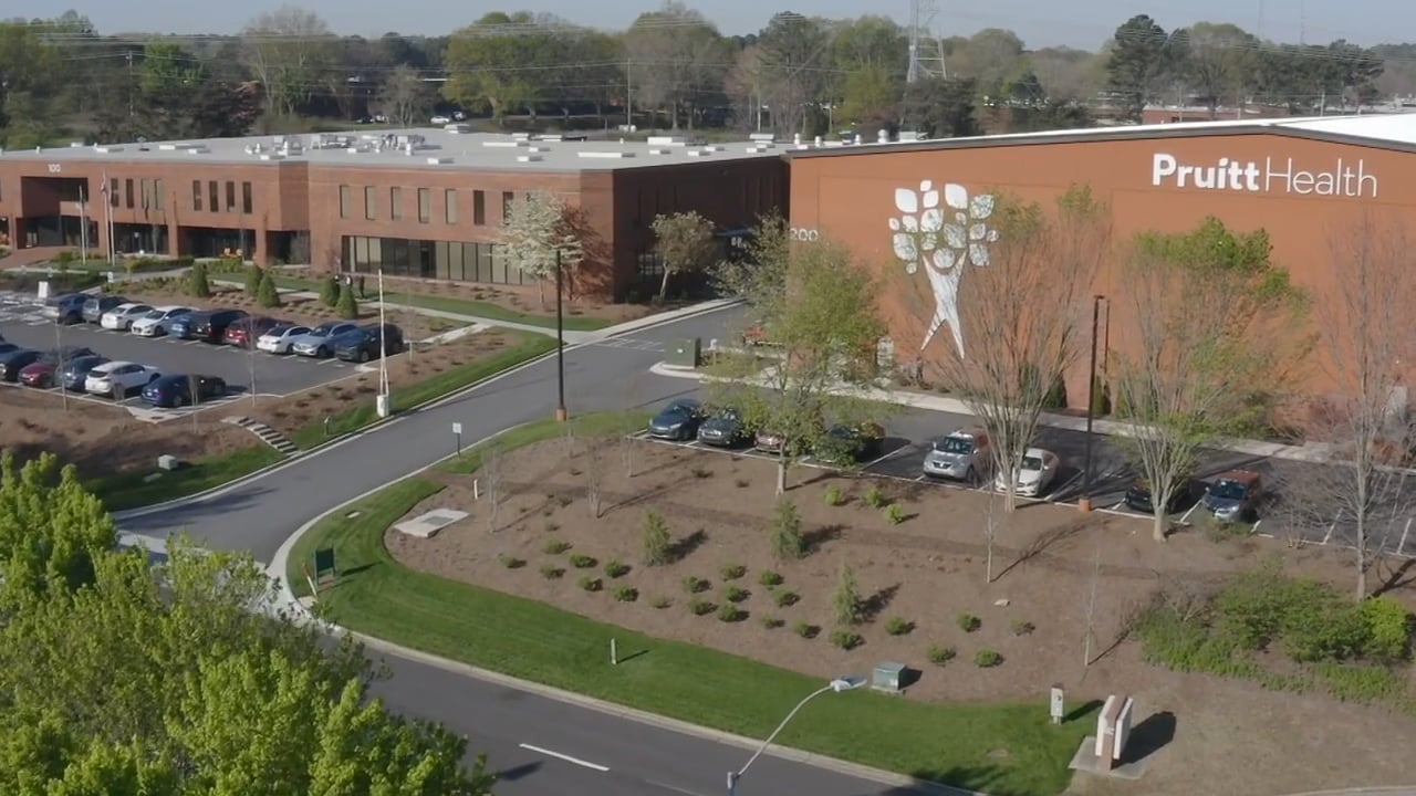 Aerial view of a large building with a parking lot and landscaped area in the foreground, surrounded by greenery.