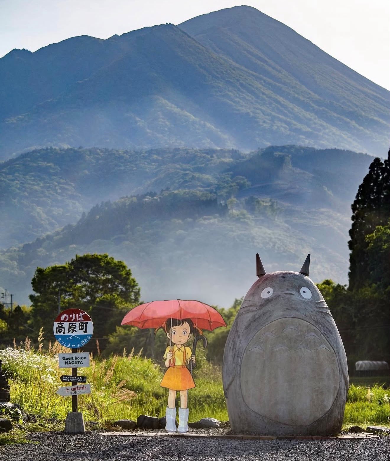 Life-size concrete Totoro sculpture and digitally illustrated Satsuki character with red umbrella at a bus stop in Takaharu, Miyazaki Prefecture, with Mount Kirishima in the background